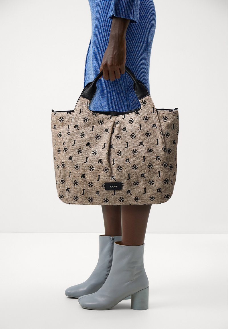 Personne en robe bleue tenant un sac fourre-tout beige à motifs avec des poignées noires et des bottines grises à talons blocs.