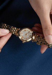 Orologio da polso in tonalità oro con un quadrante rotondo in madreperla bianca, numeri romani e un bracciale texturizzato con link lucidi alternati.