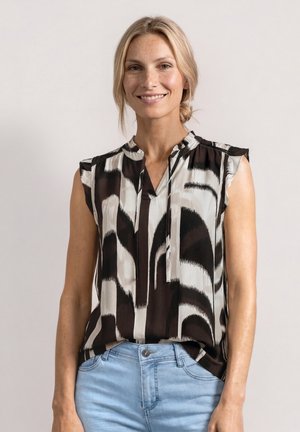 Femme souriante aux cheveux blonds attachés en queue-de-cheval basse, portant un chemisier sans manches à motifs noir et blanc et un jean bleu clair, debout devant un fond uni.