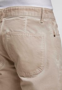 Lähikuva vaaleista housuista, joissa on suur seljatasku ja nähtavad õmblused, kantud valge särk sisse pandud.