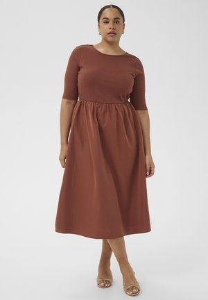 Kvinde iført en rustfarvet, mellemlang kjole med albuelange ærmer og en samlet talje, stående barfodet på ét ben mod en ensfarvet lys baggrund.