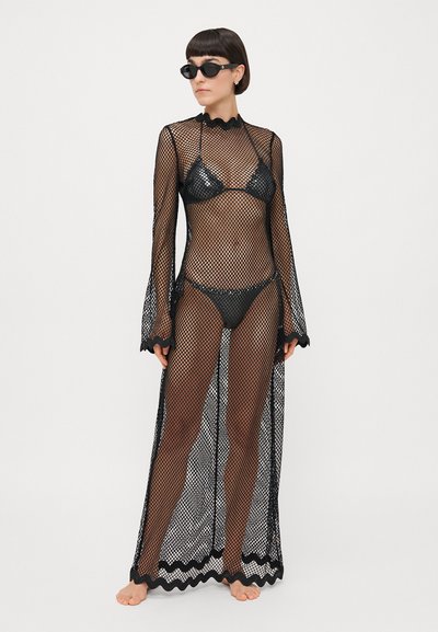 Couverture en résille noire à manches longues et bords en scallop, superposée sur un bikini noir. Texture transparente et design ouvert avec une silhouette fluide.