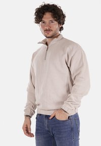 Felpa beige con zip, maniche lunghe, polsini a coste e superficie testurizzata. Indossata con jeans in denim blu.