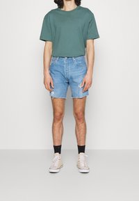 Green cotton t-shirt, blue denim shorts with frayed hem and distressed detailing, paired with beige sneakers and black socks.
