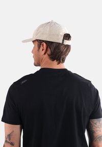 Casquette en velours côtelé beige avec visière incurvée et strap ajustable, portée par une personne en t-shirt noir arborant un logo sur l'épaule.