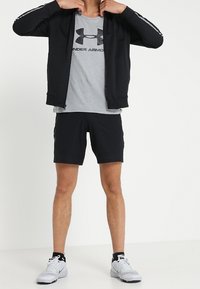 Schwarze Sportjacke mit weißen Streifen, graues Unterziehshirt von Under Armour, schwarze Shorts und graue Sportschuhe mit gelben Akzenten.