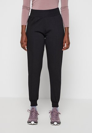 Femme portant un pantalon de jogging noir ajusté, un haut à manches longues mauve, des chaussettes bleu clair et des chaussures de sport violettes, debout devant un fond uni.