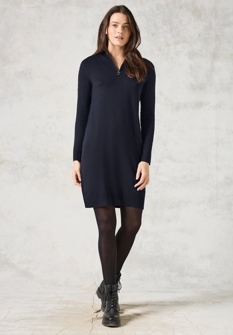 Vestito a maglia blu navy con maniche lunghe e colletto con zip, caratterizzato da un orlo a costine. Abbinato a stivaletti neri alla caviglia, su uno sfondo chiaro texturizzato.