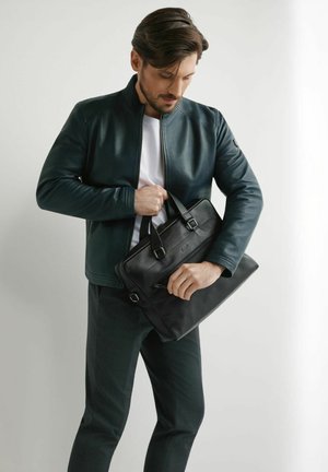 Mężczyzna w ciemnej kurtce i spodniach trzymający i otwierający czarną skórzaną teczkę na prostym jasnym tle.