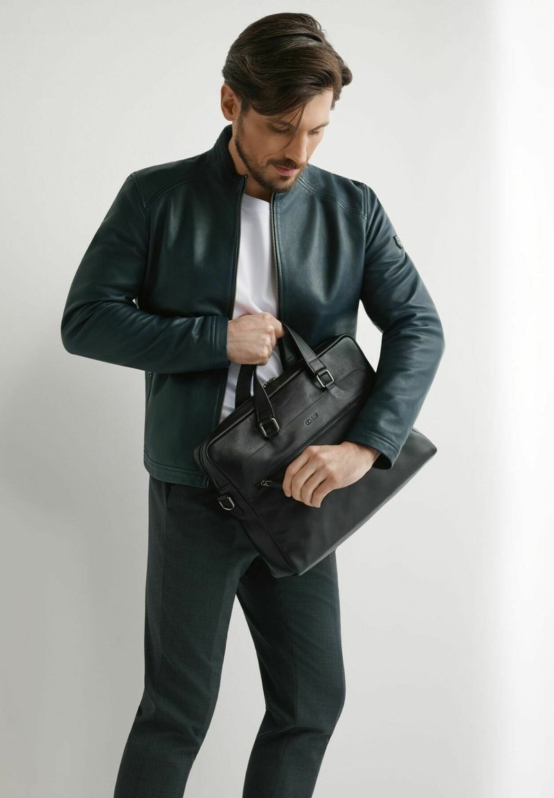 Mężczyzna w ciemnej kurtce i spodniach trzymający i otwierający czarną skórzaną teczkę na prostym jasnym tle.