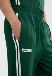 Zielone spodnie sportowe z elastycznym paskiem w talii, kieszeniami bocznymi, akcentami w białe pasy oraz małym logo "ellesse" z przodu. Gładka faktura materiału.