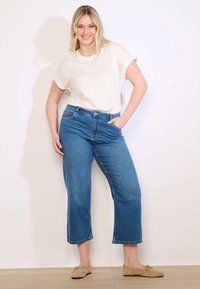 Naine seisab valge seina ääres, kandes valget t-särki, siniseid lühendatud teksaseid ja beeži moccasin-tüüpi kingi, naeratades ja hoides ühte kätt taskus.