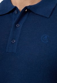 Marinblå pikétröja med ett mjukt stickat tyg, tre knappslå och en tonad broderad logotyp på bröstet.