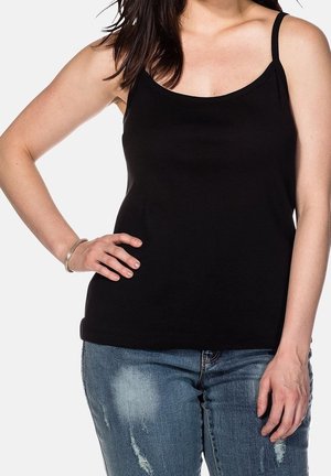 Femme portant un haut noir sans manches à fines bretelles et un jean bleu déchiré, une main sur la hanche, devant un fond uni.
