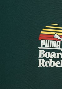 Camiseta de manga corta verde oscuro con un estampado gráfico en blanco y amarillo que incluye el texto "PUMA Board Rebel" y un diseño de rayos de sol estilizado.