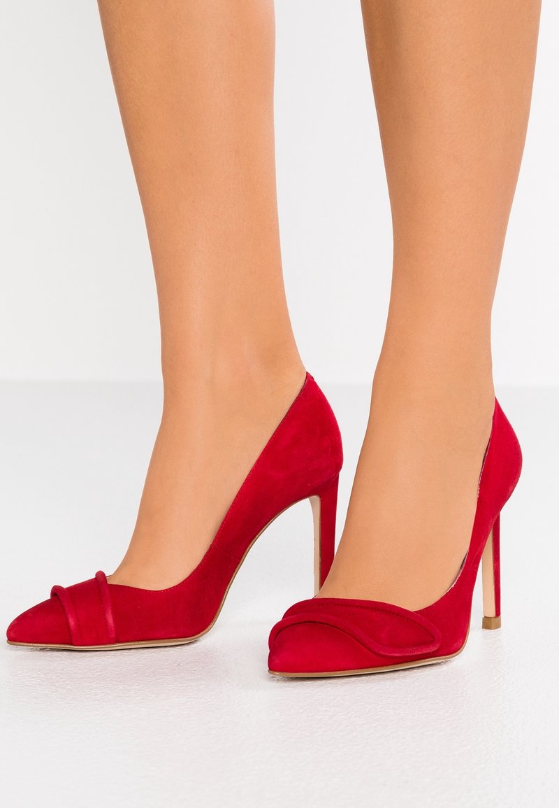 Femme portant des escarpins rouges en daim à talons hauts avec un bout pointu et une bride décorative à l'avant, debout sur une surface blanche.