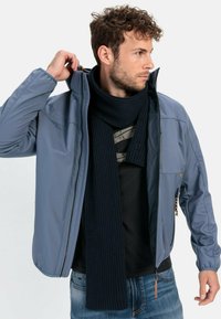 Blå softshell-jakke med høj krave, lynlås, brystlomme, parret med et mørkt ribbet tørklæde, båret over en sort skjorte og jeans.