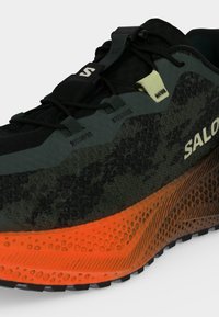 Μαύρο και πράσινο αθλητικό παπούτσι Salomon με υφή πορτοκαλί σόλας και σύστημα γρήγορου δέσιμου σε λευκό φόντο.