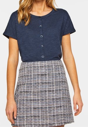 Femme portant un haut bleu marine à manches courtes boutonné, rentré dans une jupe taille haute à carreaux gris et blancs.