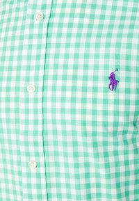 Zelená a bílá károvaná košile na knoflíky z látky s hladkou texturou, s fialovým vyšitým logem na levé straně hrudi.