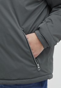 Grå polstret jakke med glidelås-lomme, glatt tekstur, og design med tettsittende mansjetter. Jakken har en moderne fasong med sømdetaljer.