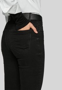 Black denim jeans with a fitted design, featuring a back pocket and a black leather belt. The fabric has a smooth texture.