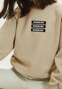 Felpa beige con tre toppe nere ricamate con la scritta "WHENEVER". Tessuto morbido, vestibilità rilassata, polsini e orlo a costine, design minimalista.