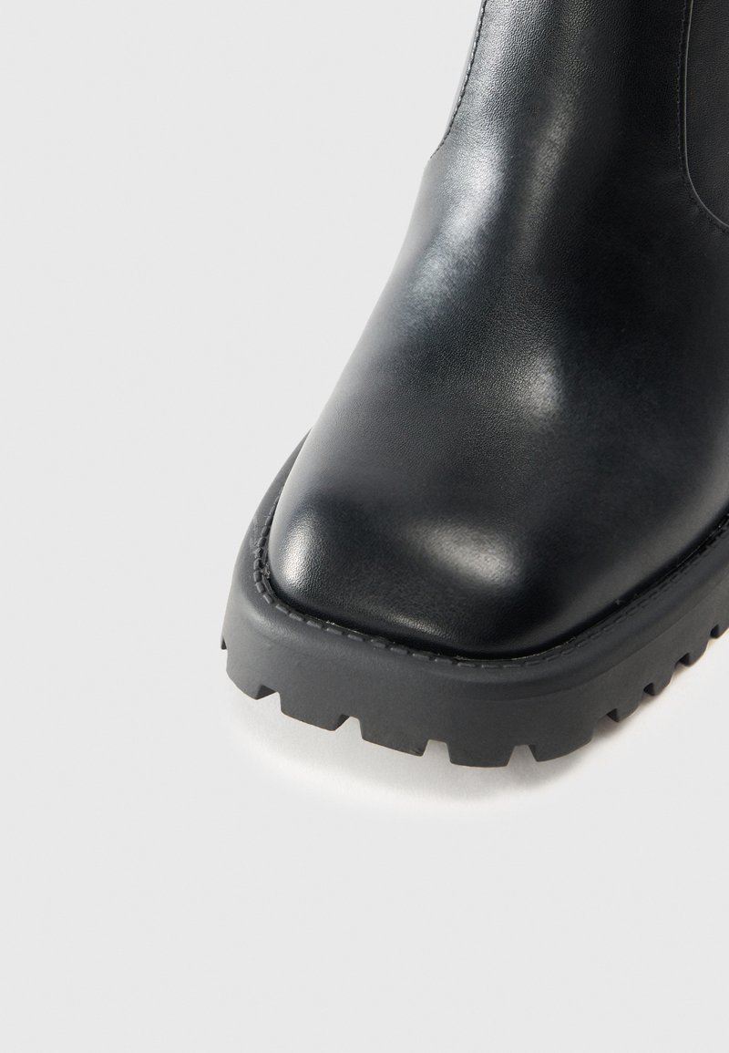 Bottine en cuir noir avec une finition lisse, bout pointu et semelle en caoutchouc épaisse dotée d'un motif de bande de roulement dentelé.