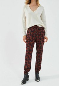 Cremfarbener V-Ausschnitt-Pullover und schwarze Hose mit floralen roten und grünen Mustern, mit elastischem Bund und eng anliegenden Bündchen, kombiniert mit schwarzen Knöchelstiefeln.
