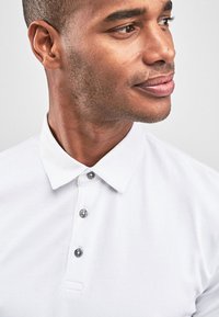 Hvid polo skjorte med en klassisk krave, der har tre grå knapper ved halsen. Glat stofstruktur, minimalistisk design.