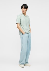 Νεαρός άνδρας με τα χέρια στις τσέπες, φοράει ανοιχτό πράσινο ριγέ κοντομάνικο πουκάμισο, ανοιχτό μπλε τζιν παντελόνι με φαρδιά μπατζάκια και λευκά αθλητικά παπούτσια, όρθιος.