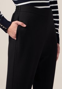 Svarta, stretchy tygbyxor med en slät yta, som har sidofickor och resårmidja för komfort. Nära bild med handdetalj.