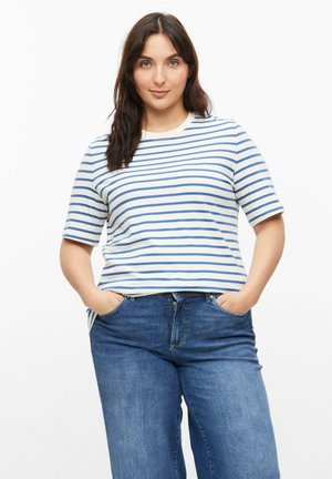 Frau mit dunklem Haar, die ein weiß-blau gestreiftes kurzärmeliges Hemd und blaue Jeans trägt, steht mit den Händen in den Taschen vor weißem Hintergrund.