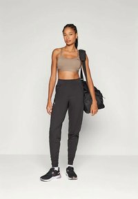 Sportliches Outfit mit einem hellbraunen Crop-Top, schwarzen, tapered Jogginghosen mit Logo und schwarzen Sneakers mit pinken Akzenten. Trägt eine schwarze Sporttasche.