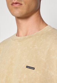 Beige színű pamut póló, textúrázott felülettel, kerek nyakkal és a bal oldalon található kis fekete "UNDER ARMOUR" logóval.