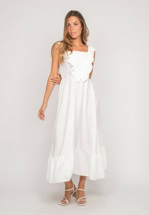 Femme portant une robe longue blanche sans manches avec un col et un ourlet volantés, associée à des talons blancs à brides, se tenant devant un fond uni.