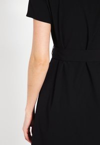 Musta mekko, jossa on lyhyet hihat ja vyötäröllä kireä kangashihna. Sujuva materiaali, jossa on yksinkertainen ja selkeä muotoilu.