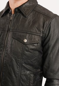 Černá kožená bunda s předním zipem, zapínacími klopa kapsami a klasickým límcem. Obsahuje texturovaný materiál a viditelné detaily stehu.