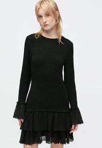 Vestido negro con mangas largas, tejido texturizado, dobladillo con volantes y ribete de encaje en las mangas, creando una silueta en capas.