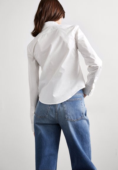Cămașă albă cu guler și hem curbat, asociată cu blugi din denim de un albastru deschis, largi. Blugii au buzunare din spate standard.