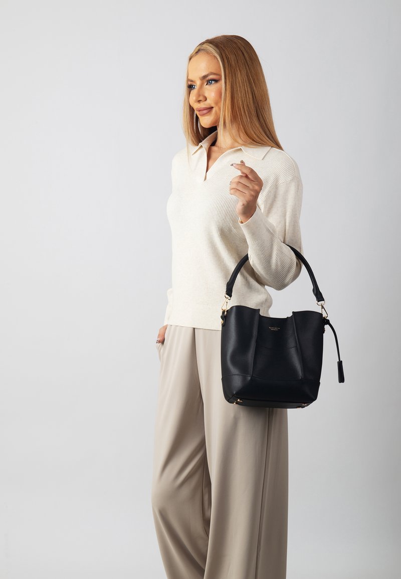Black leather bucket bag with a gold accent, featuring a curved handle and a smooth texture. Worn by a person in light-colored clothing.