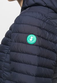 Tummansininen tilkkutakki, jonka olalla on vihreä ympyrän muotoinen "Save the Duck" -logopatch, jossa on valkoinen ankka kierrätysnauhojen kanssa.