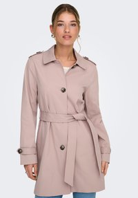 Trench-coat rose clair avec une taille ceinturée, des boutons sur les poignets, et un col classique. Fabriqué en tissu lisse et léger.
