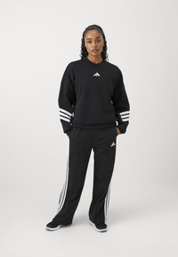 Sweatshirt noir avec logo Adidas blanc et trois bandes sur les manches, associé à un pantalon noir avec des bandes blanches sur les côtés.
