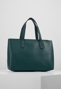 Tumma vihreä teksturoitu nahkatoimikassi, jossa on kaksi kahvaa, seisomassa pystyasennossa valkoisella jalustalla tasaisen harmaan taustan edessä.