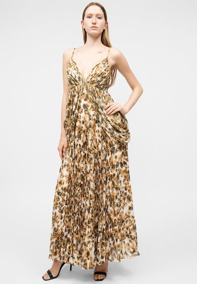 Γυναίκα όρθια με το χέρι στη μέση, φορώντας μανίκια, μακρύ φόρεμα με πλισέ σχέδιο λεοπάρ και μαύρες ψηλοτάκουνες σανδάλες.