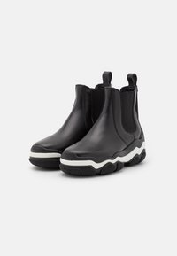 Par de botas de cuero negro hasta el tobillo con acentos blancos, paneles laterales elásticos y suelas de goma texturizada sobre un fondo blanco.
