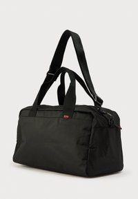 Μαύρη τσάντα duffel από νάιλον με ορθογώνιο σχήμα. Διαθέτει διπλές χειρολαβές, ρυθμιζόμενο ιμάντα και κλεισίματα με φερμουάρ στο πάνω μέρος.