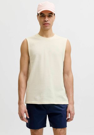 Homme portant un débardeur beige clair, un short bleu marine et une casquette de baseball rose pâle, debout devant un fond uni.