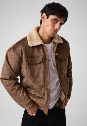 Junger Mann trägt eine braune Wildlederjacke mit einem beigen Lammfellkragen und einem weißen T-Shirt, posiert mit den Händen in den Taschen.